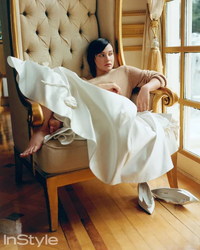 Cailee Spaeny reclining barefoot in an armchair beside a window.