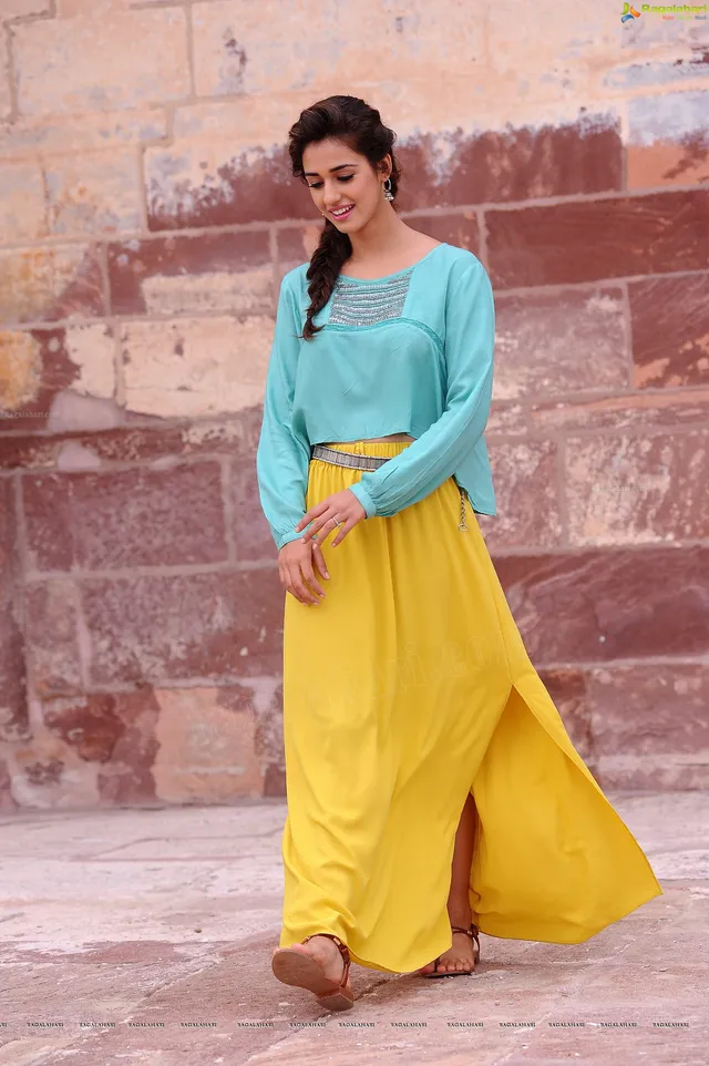 Disha Patani standing in flat sandals with yellow trousers and a turquoise blouse.
