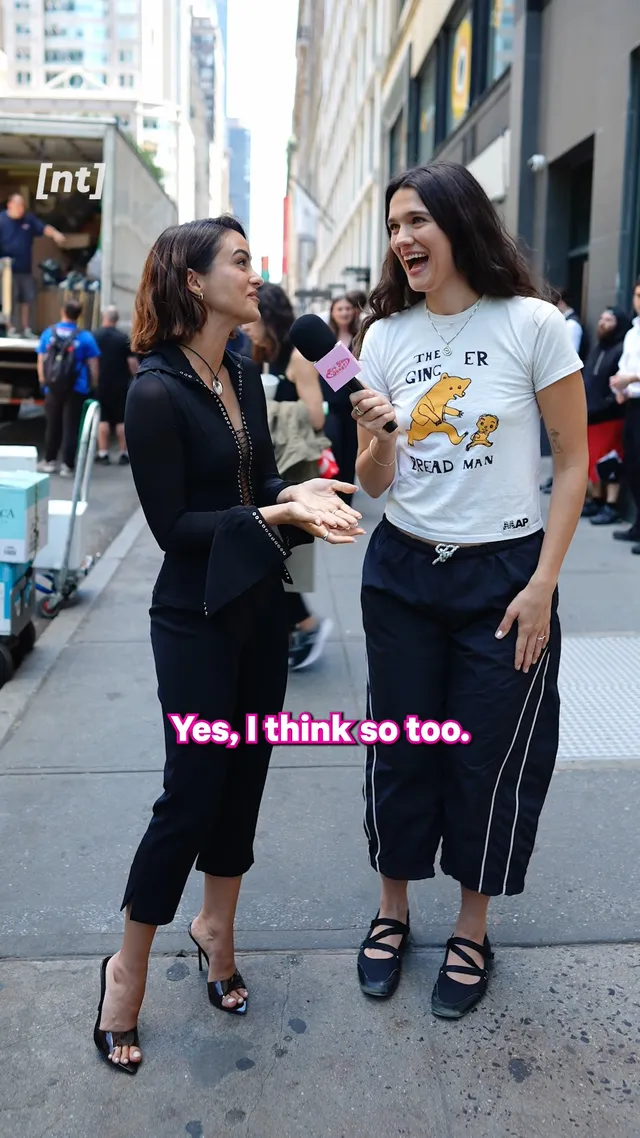 Camila Mendes giving an outdoor interview in a black outfit and heels.