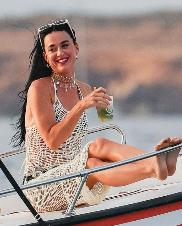 Katy Perry sitting barefoot aboard a boat with a drink.