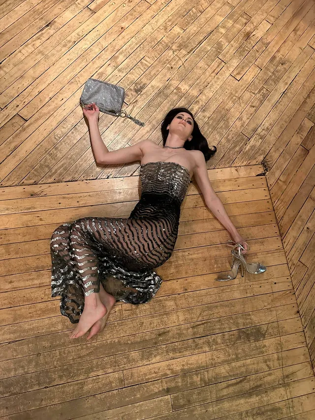 Alexandra Daddario lying on a wooden floor in a dark lace dress with her bare feet visible.