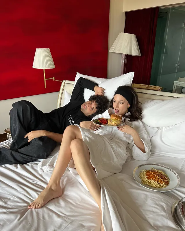 Úrsula Corberó reclining barefoot in a pale dress beside a man on a bed.