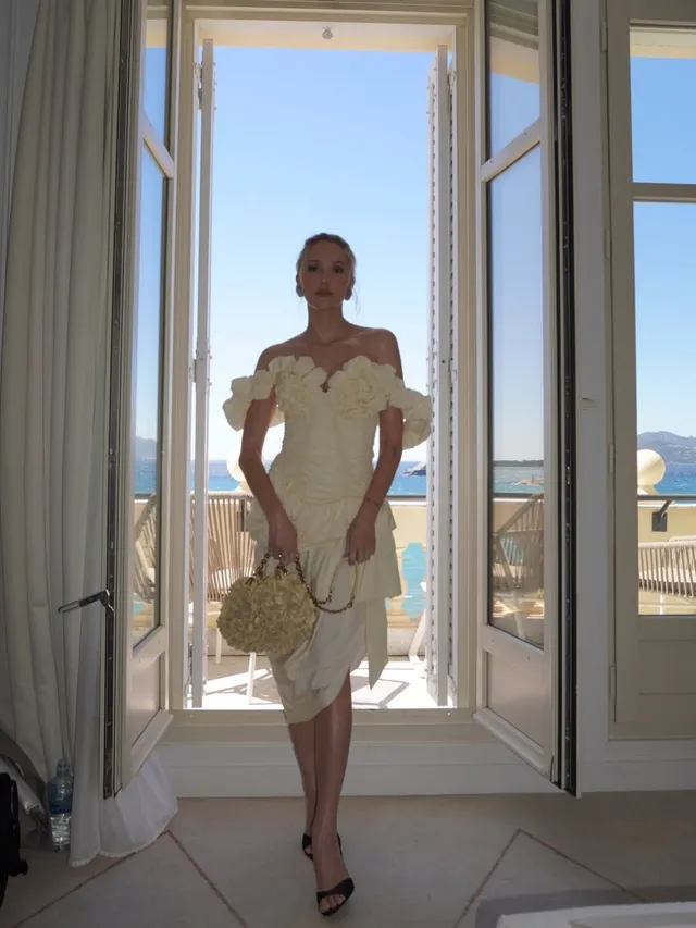 Maika Monroe wearing a pale dress and black heels by balcony doors.