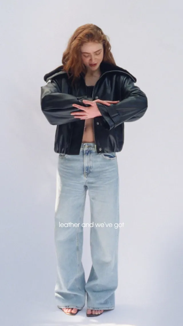 Sadie Sink standing barefoot in jeans and a leather jacket.
