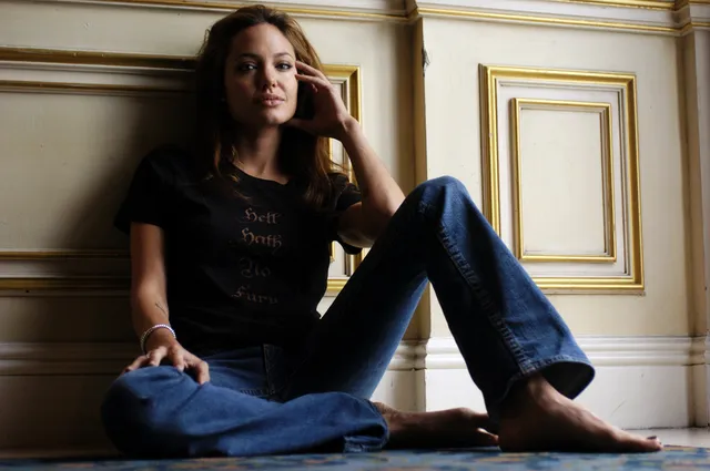 Angelina Jolie seated barefoot against a wall in a dark shirt and jeans.