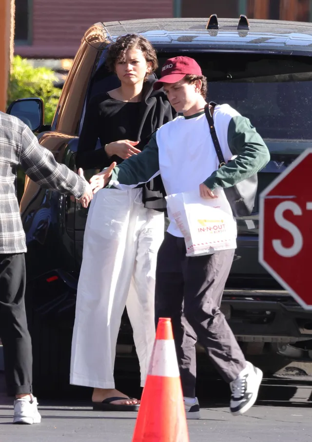 Zendaya walking with other people in white trousers and flat sandals.