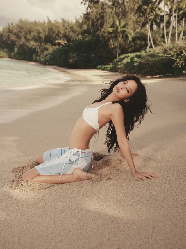 Jennie Kim kneeling barefoot on a beach in a white top and a pale blue skirt.