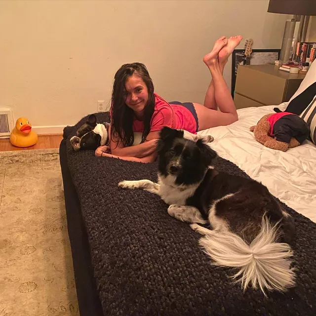 Nina Dobrev lying on a bed beside dogs with bare feet raised.