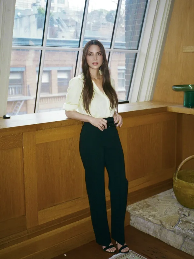 Olivia Munn posing beside a window in trousers and strappy heels.