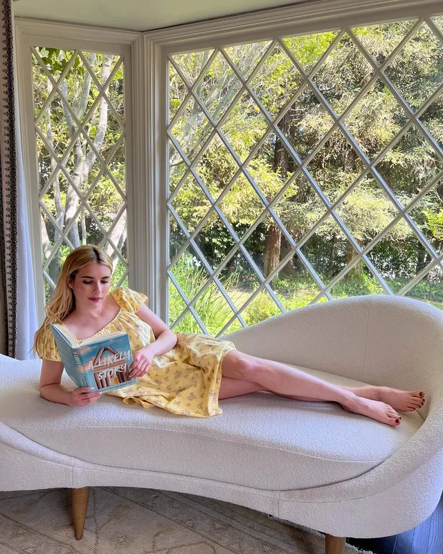 Emma Roberts reading barefoot on a chaise beside a window in a yellow dress.