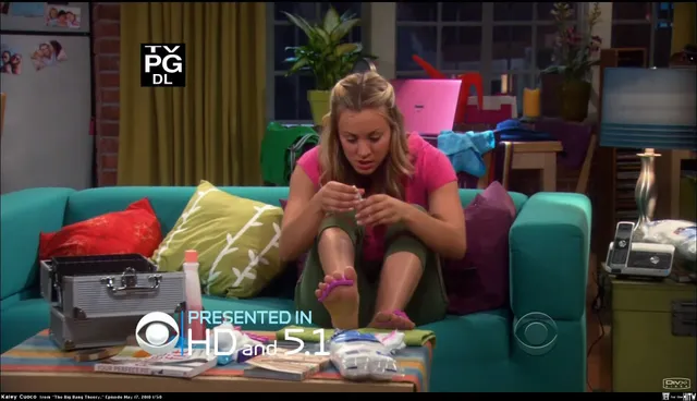 Kaley Cuoco leaning forward on a turquoise sofa with her bare feet visible in a television scene.