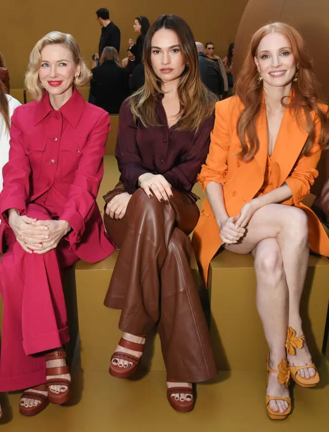 Lily James sitting between two guests in brown trousers and heels.