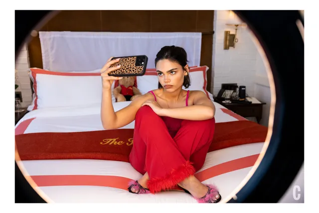 Ariana Greenblatt sitting in a red outfit and flat sandals on a bed.
