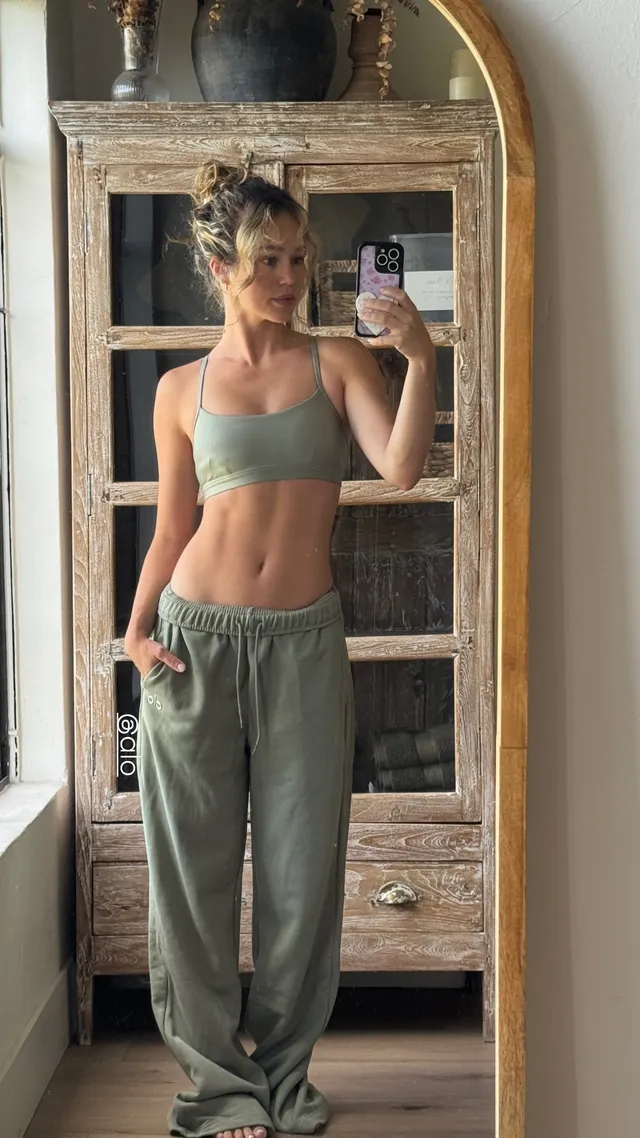 Brec Bassinger taking a mirror selfie in green trousers and a cropped top with bare feet visible.