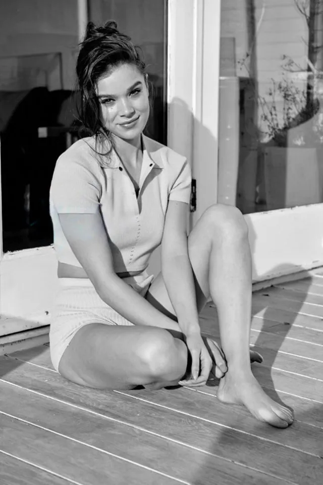 Hailee Steinfeld sitting barefoot on a wooden floor in a black and white portrait.