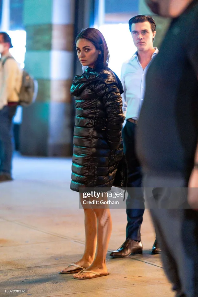 Mila Kunis standing barefoot on a pavement in a black dress.
