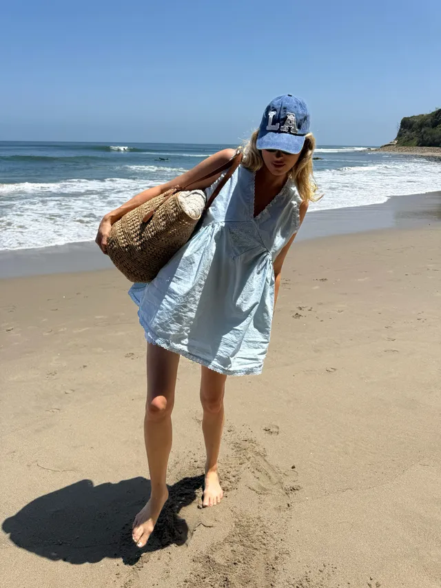 Elsa Hosk standing barefoot on a beach in a blue shirt and cap.