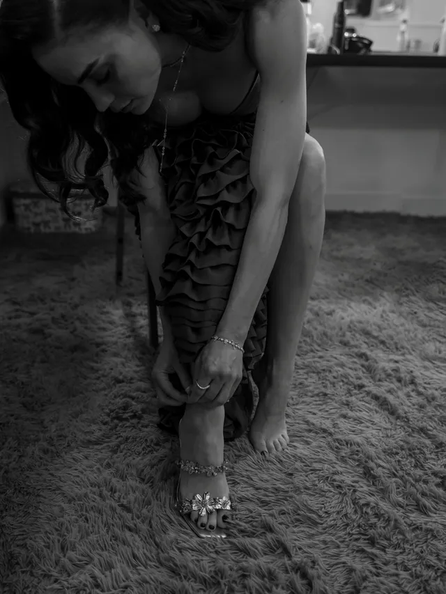 Melissa Barrera adjusting a heeled sandal in a monochrome portrait.