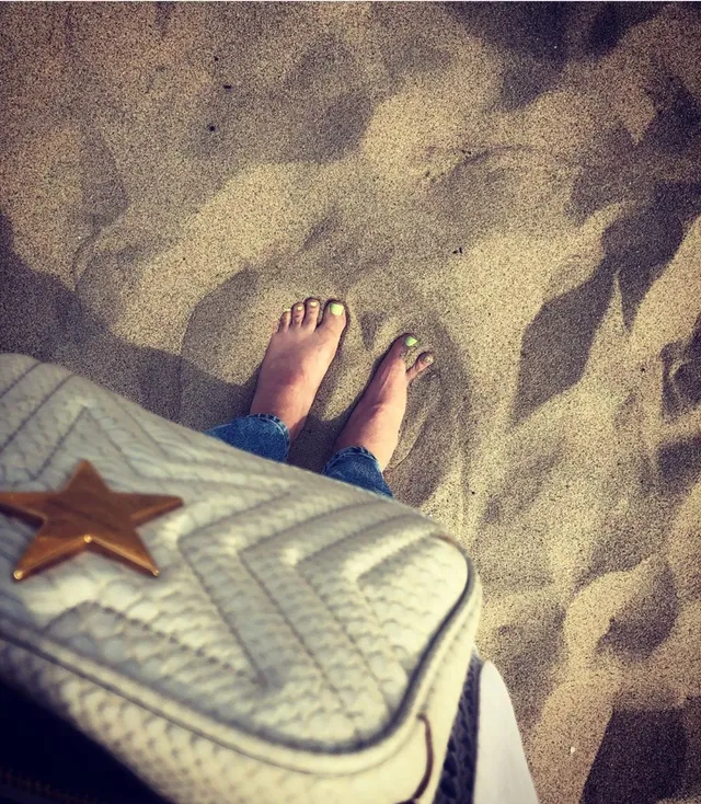 Elisha Cuthbert standing barefoot in sand beside a bag.
