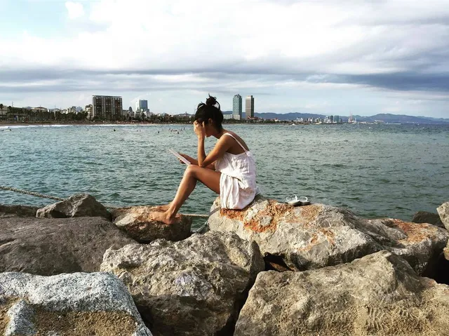 Anya Chalotra sitting barefoot on rocks beside the sea.