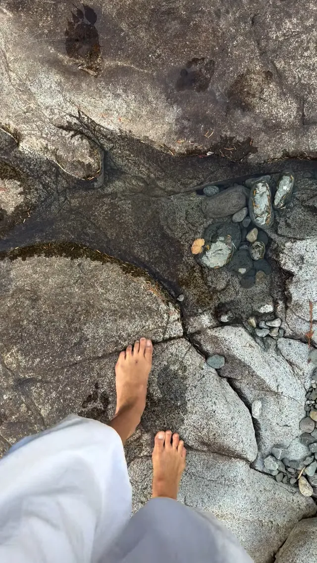 Bella Hadid standing barefoot on rocks beside water.