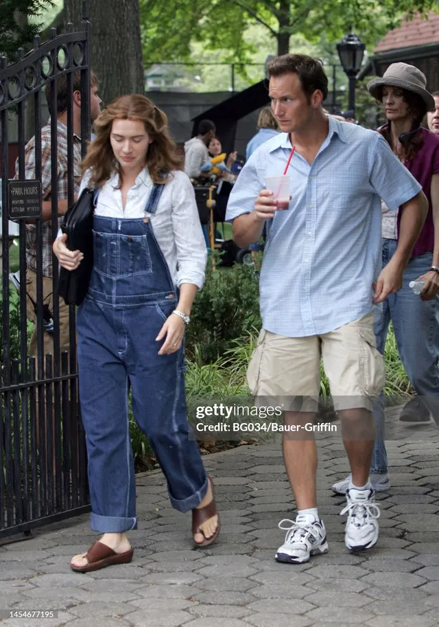 Kate Winslet walking outdoors in denim overalls and sandals beside a companion.