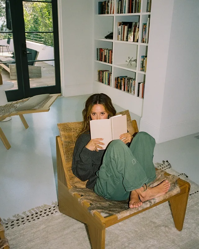 Ashley Tisdale sitting in a chair with her bare feet raised while reading.