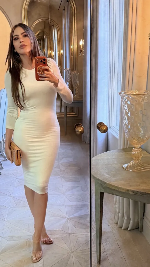Sofía Vergara posing for a mirror photograph in a white dress and transparent heels.