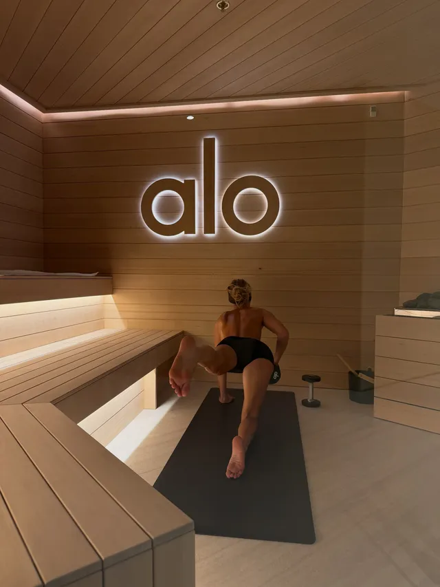 Bella Hadid stretching barefoot on a mat in a wood-paneled room.
