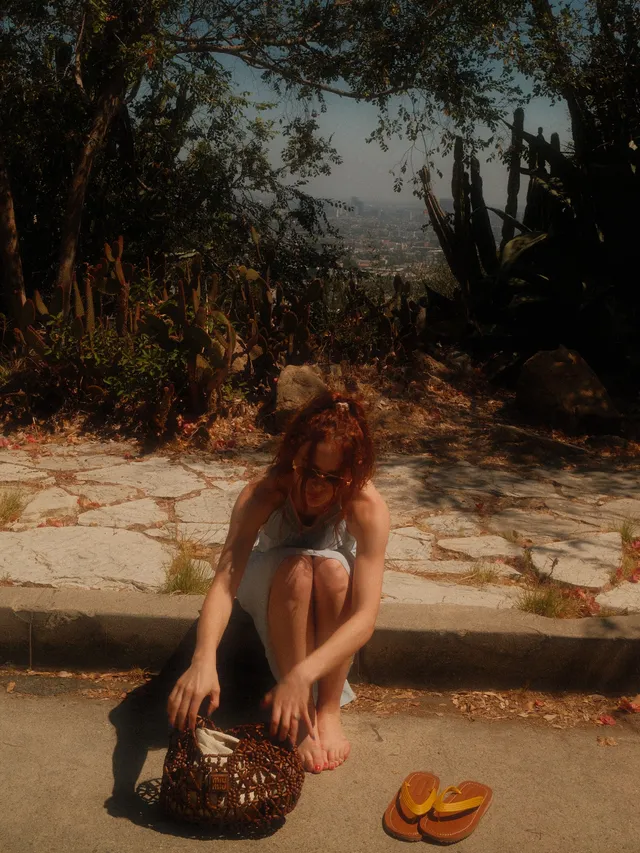 Joey King sitting barefoot outdoors beside a picnic basket.