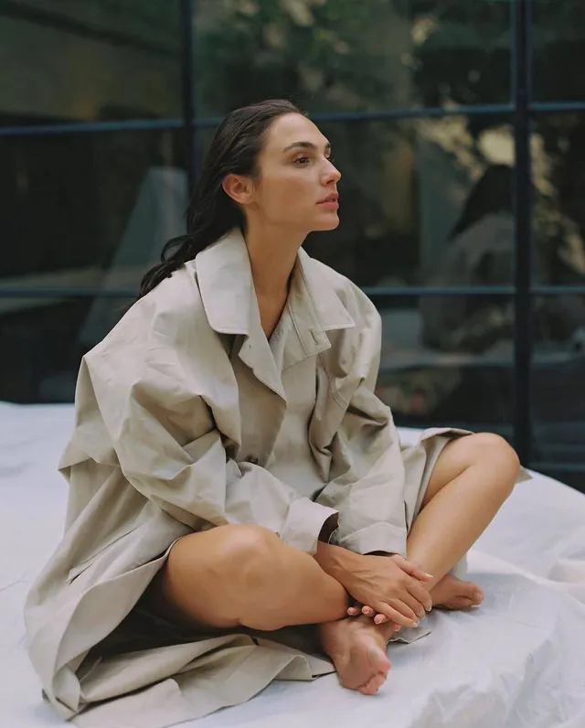 Gal Gadot sitting barefoot on a bed wearing a light coat.
