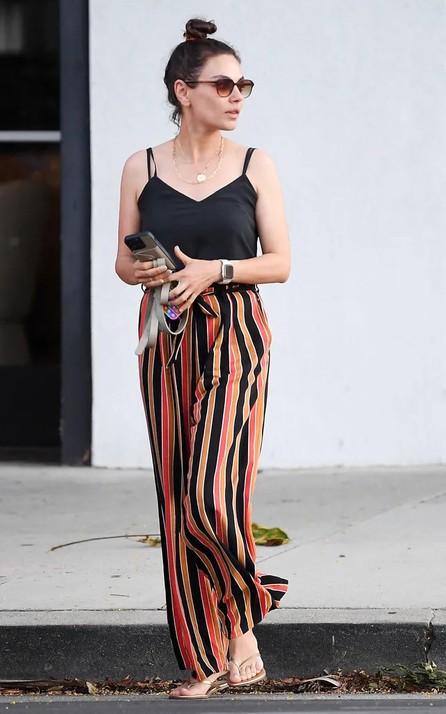 Mila Kunis standing outdoors in striped trousers and flip-flops.