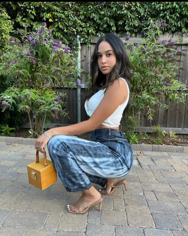 Emily Tosta crouching outdoors in jeans and clear heels.