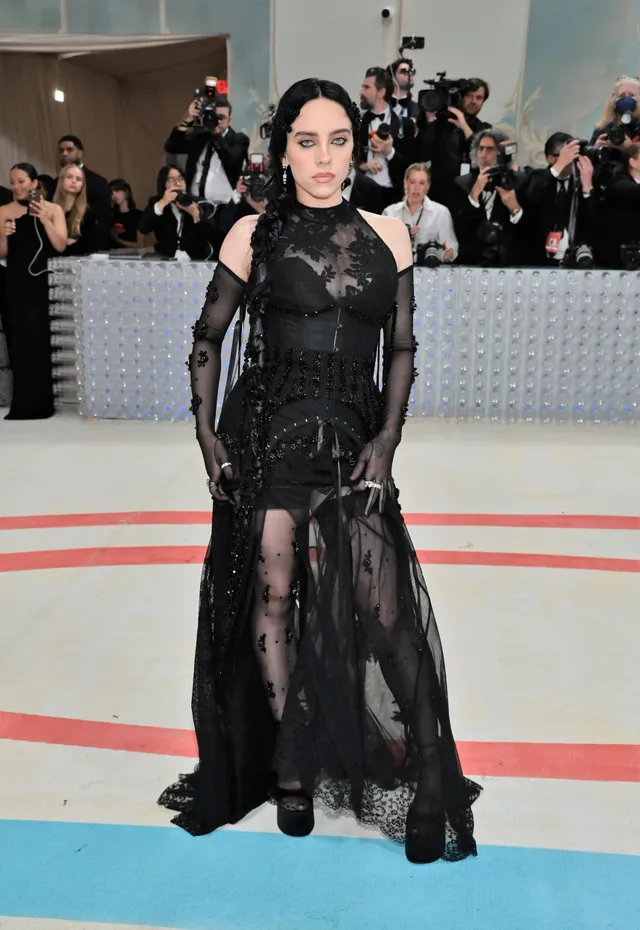Billie Eilish posing at an event in a long black dress and platform heels.