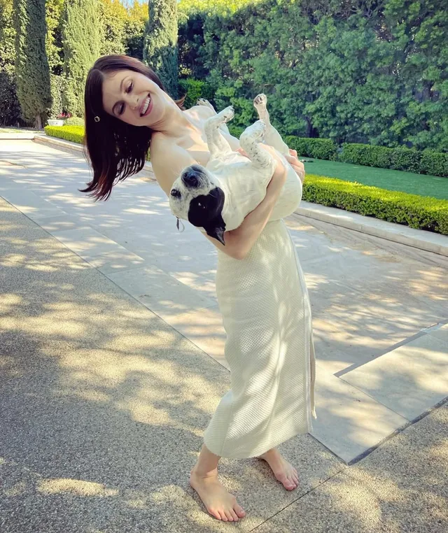 Alexandra Daddario standing barefoot outdoors in a white dress while holding a dog.