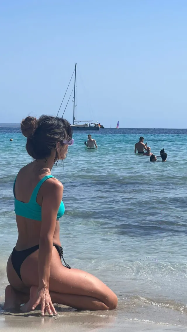 María Pedraza kneeling barefoot in shallow sea water in a bikini.