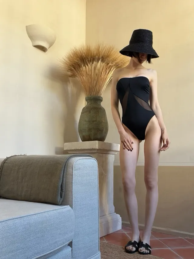 Lily Collins standing indoors in a black outfit and flat sandals.