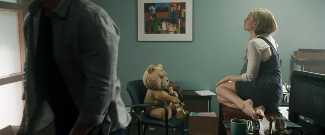 Amanda Seyfried sitting barefoot indoors beside a teddy bear in a screen image.