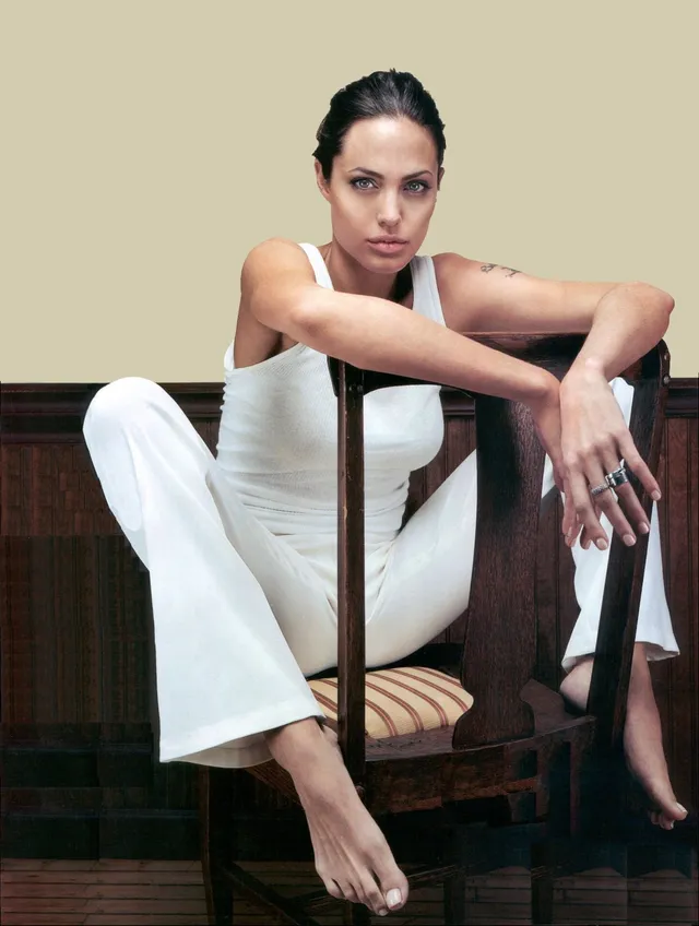 Angelina Jolie seated backwards on a wooden chair in white trousers.