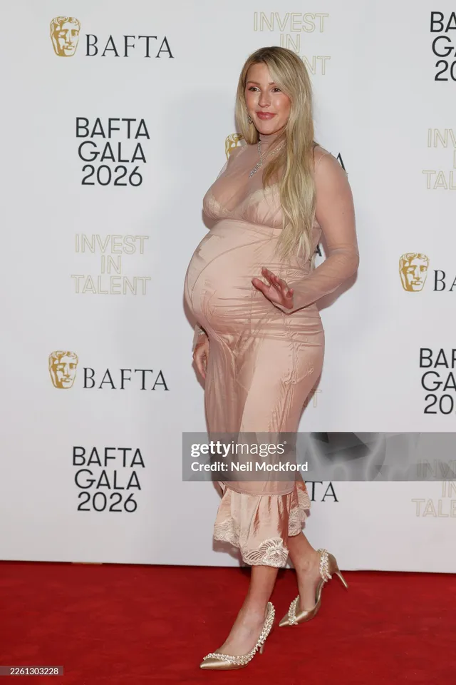Ellie Goulding posing at an event in a pale dress and white heels.