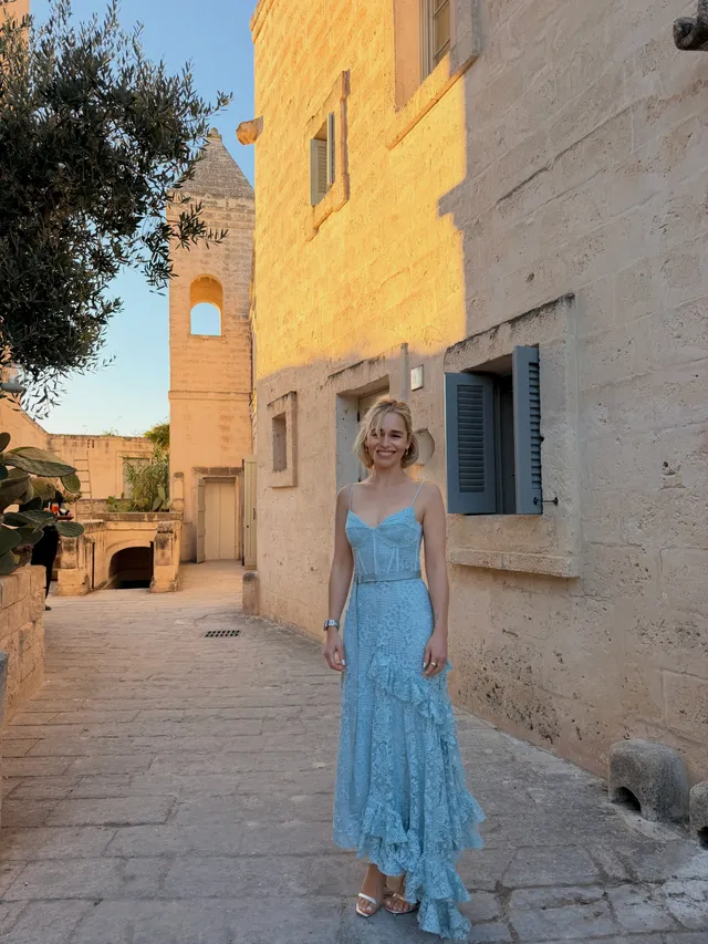 Emilia Clarke walking along a cobbled street in a blue dress and heels.