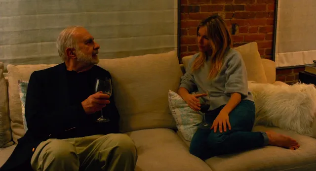 Sienna Miller sitting on a sofa beside a man in a screen scene.