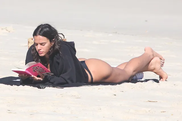 Bruna Marquezine reclining barefoot on a beach in a dark bikini while reading.