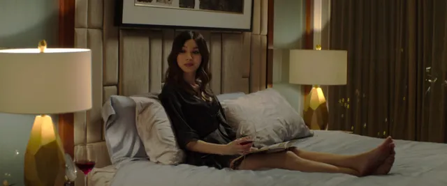 Gemma Chan sitting on a bed in a screen scene.