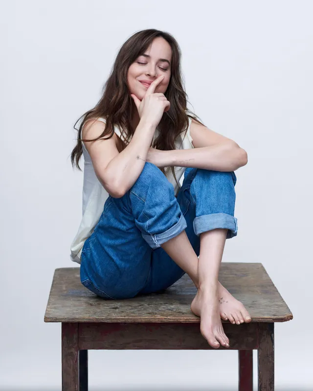 Dakota Johnson sitting barefoot on a wooden stool in jeans.