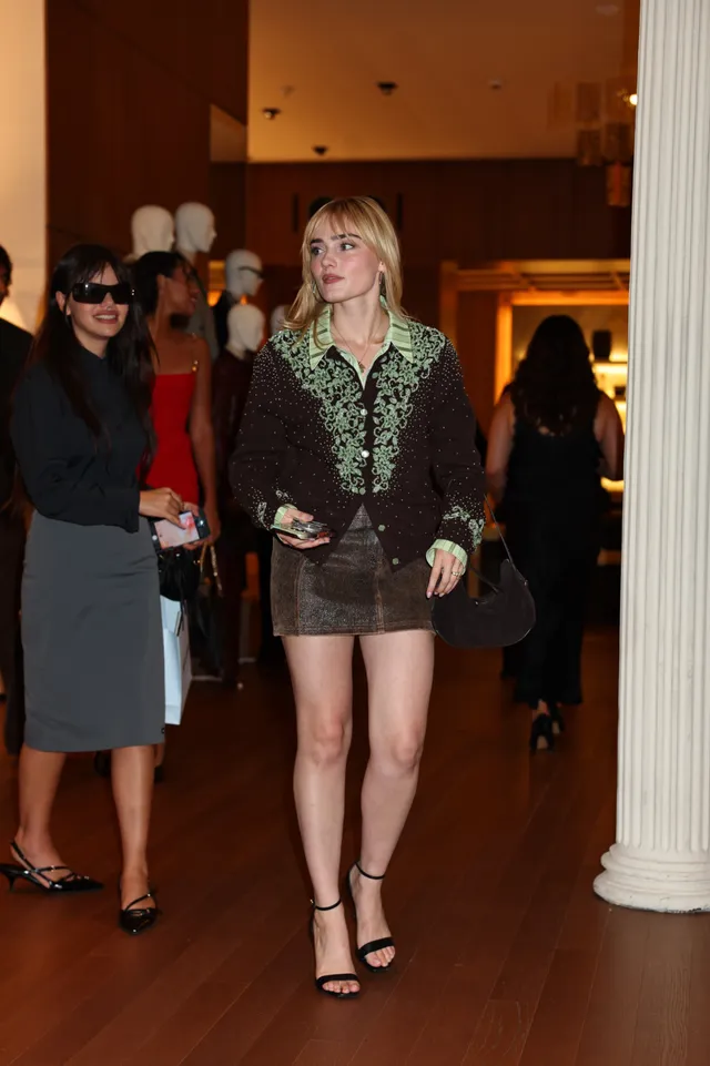 Meg Donnelly walking in a patterned blouse, brown skirt and black heels.