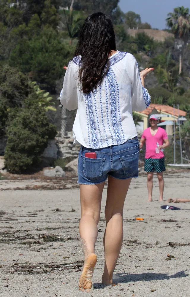Mila Kunis walking barefoot along a beach seen from behind.
