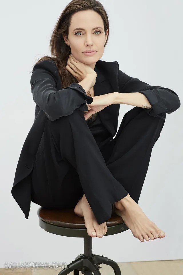 Angelina Jolie seated on a stool against a pale studio backdrop.