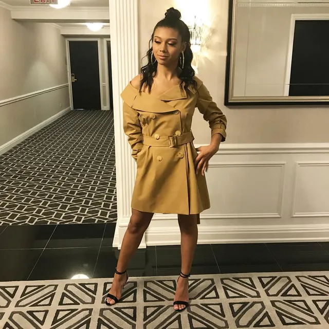 Sierra Capri posing in a yellow coat-style dress and black heels.