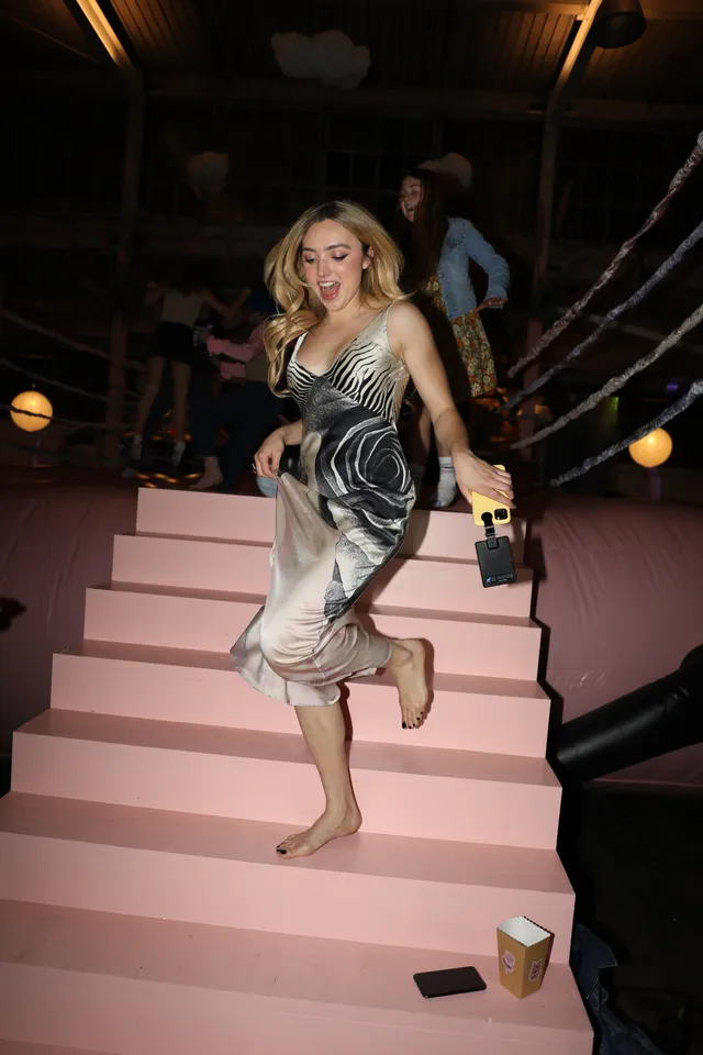 Peyton List walking down pink stairs in a dress and heels.